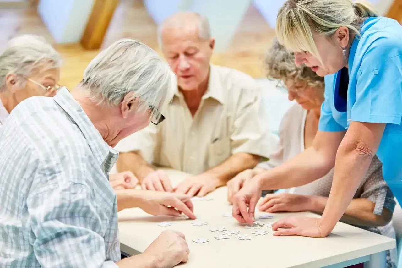 Senioren legen gemeinsam ein Puzzle, unterst&uuml;tzt durch eine Pflegekraft. Dies f&ouml;rdert die Aktivit&auml;t und soziale Interaktion, wie sie &sect;43b SGB XI vorsieht.
