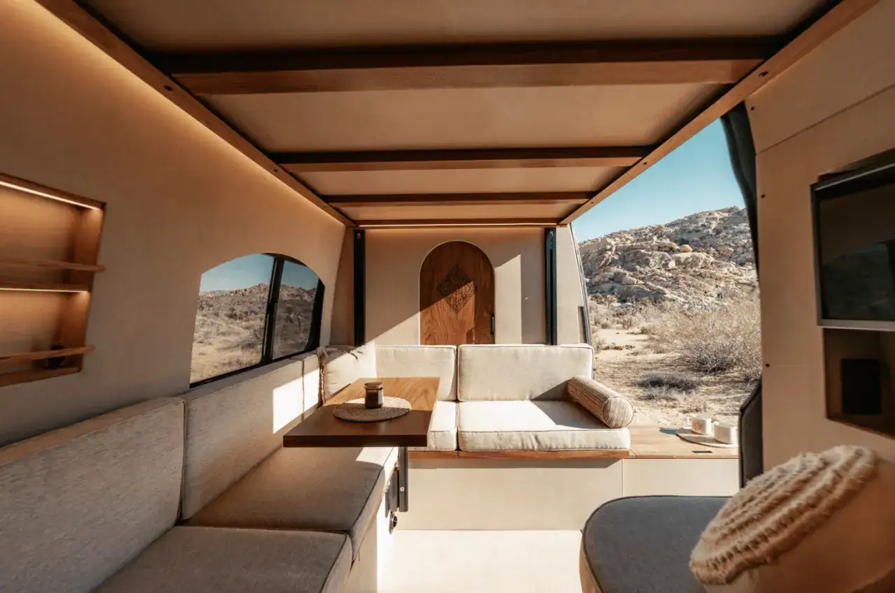 Ambiance cosy dans ce camping-car. L'intérieur offre des banquettes confortables, une table en bois et une porte décorative, le tout baigné de lumière naturelle.