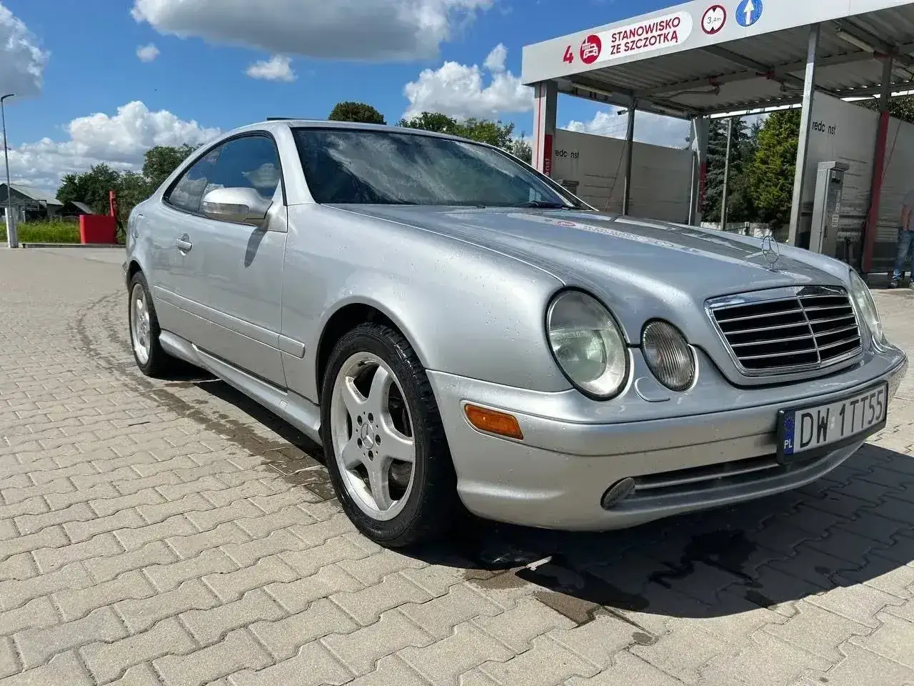 Srebrny Mercedes CLK 200 po umyciu na samoobsługowej myjni. Klasyczna sylwetka coupe na tle błękitnego nieba.