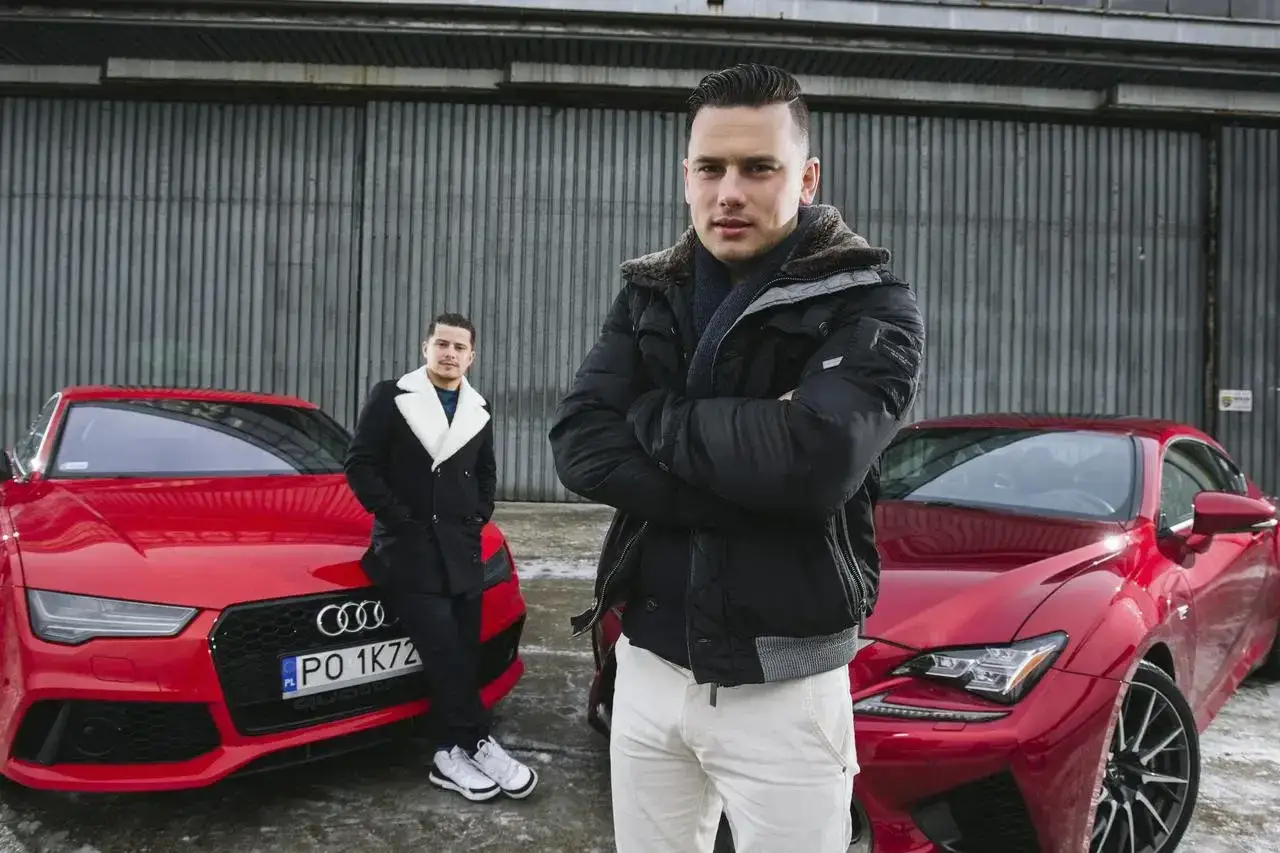 Bracia Collins pozują obok czerwonych sportowych samochodów.