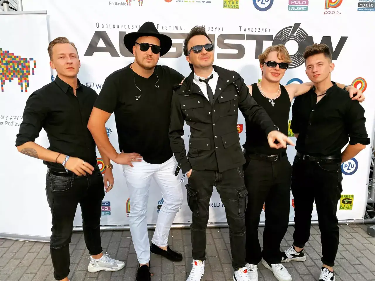 Pięciu młodych mężczyzn, prawdopodobnie zespoły disco polo, pozuje do zdjęcia na tle festiwalowego banera.