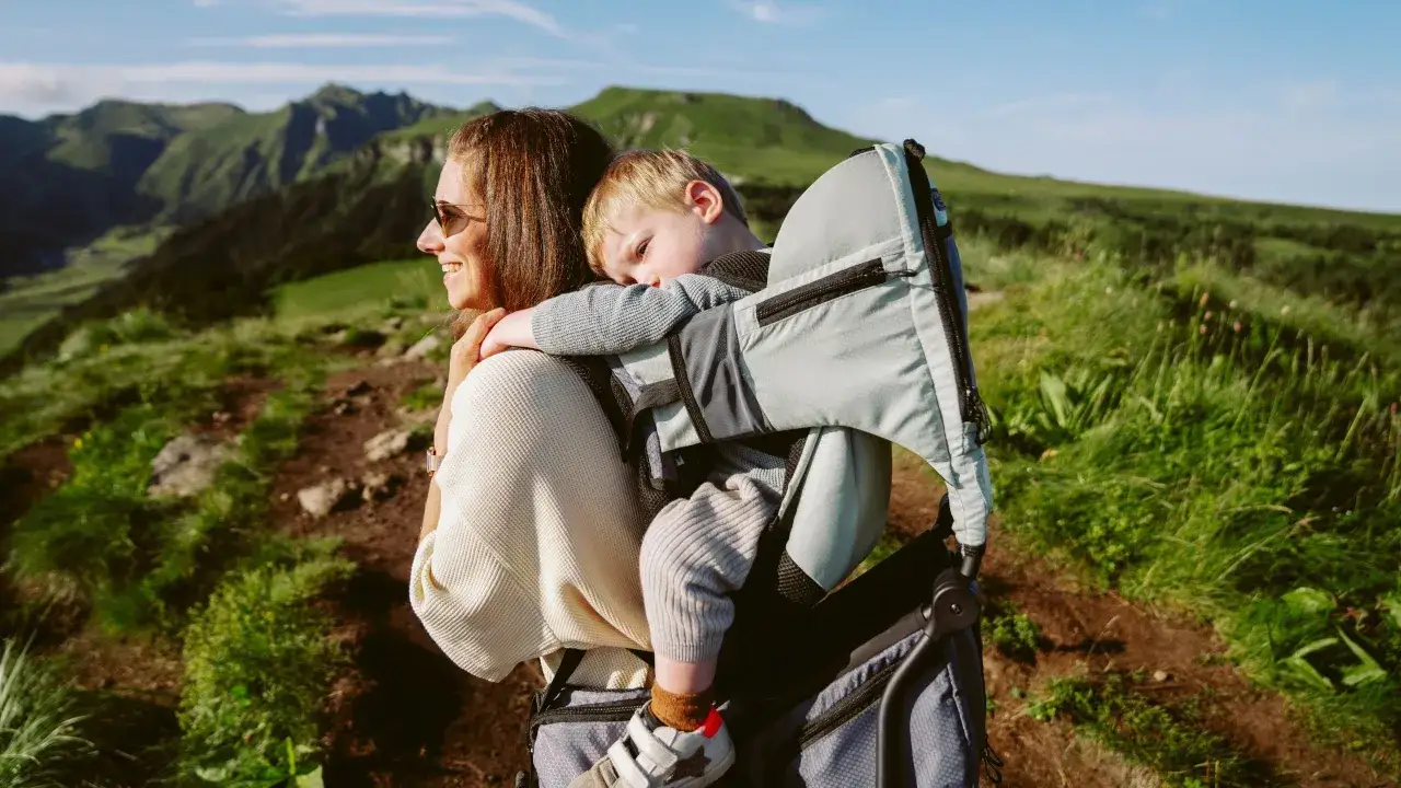 Une mère souriante porte son jeune enfant endormi dans un porte bébé rando age, sur fond de montagnes verdoyantes.