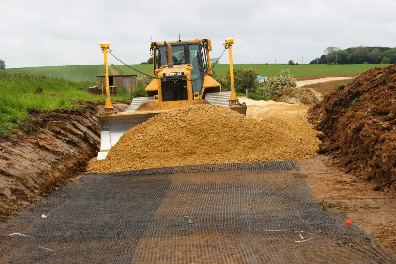 Buldożer Caterpillar przygotowuje teren pod utwardzenie drogi dojazdowej do działki, zasypując wykop żwirem.