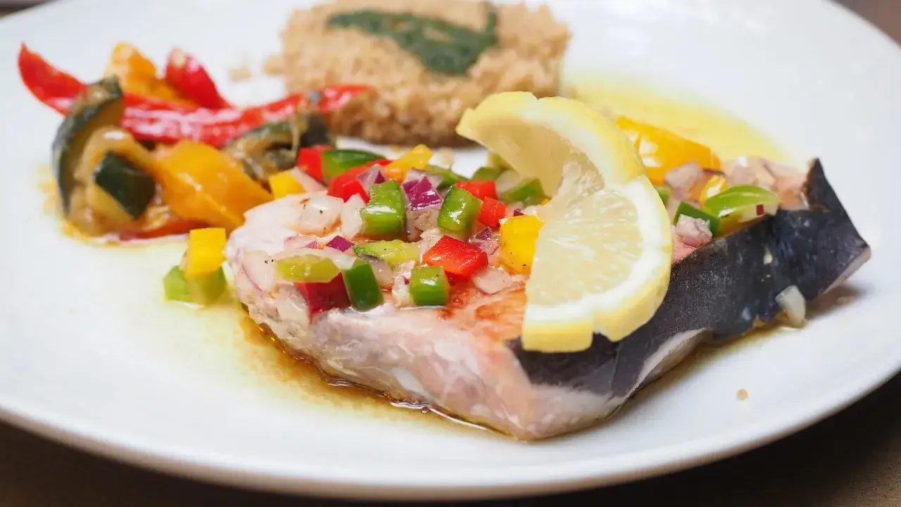 Un succulento pesce spada al forno, guarnito con un trito colorato di peperoni e cipolla, accompagnato da verdure miste e una fetta di limone.