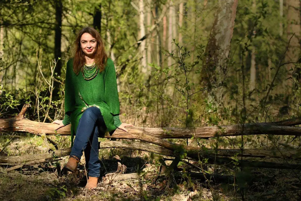 Kobieta w zielonym swetrze siedzi na powalonym drzewie w lesie. To idealne miejsce na leśny dom seniora, gdzie można cieszyć się naturą.