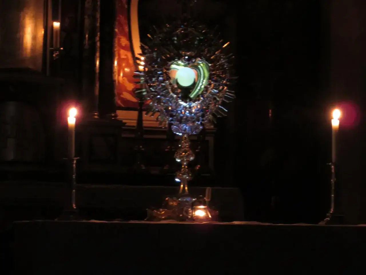 Ein gl&auml;nzendes Herz-Jesu-Monstranz im Kerzenlicht. Zwei Kerzen leuchten links und rechts, w&auml;hrend im Hintergrund eine rote Fahne zu sehen ist.