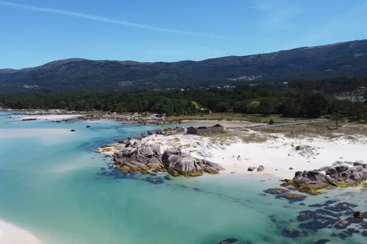 Aguas turquesas bañan la **playa Caolín**, con arena blanca, rocas y pinares al fondo.