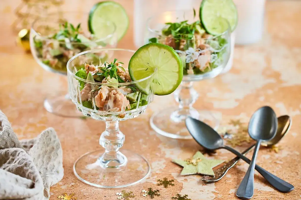 Elegante Lachs-Avocado-Vorspeise im Glas, garniert mit Limettenspalten und frischen Kr&auml;utern. Perfekt f&uuml;r festliche Anl&auml;sse.