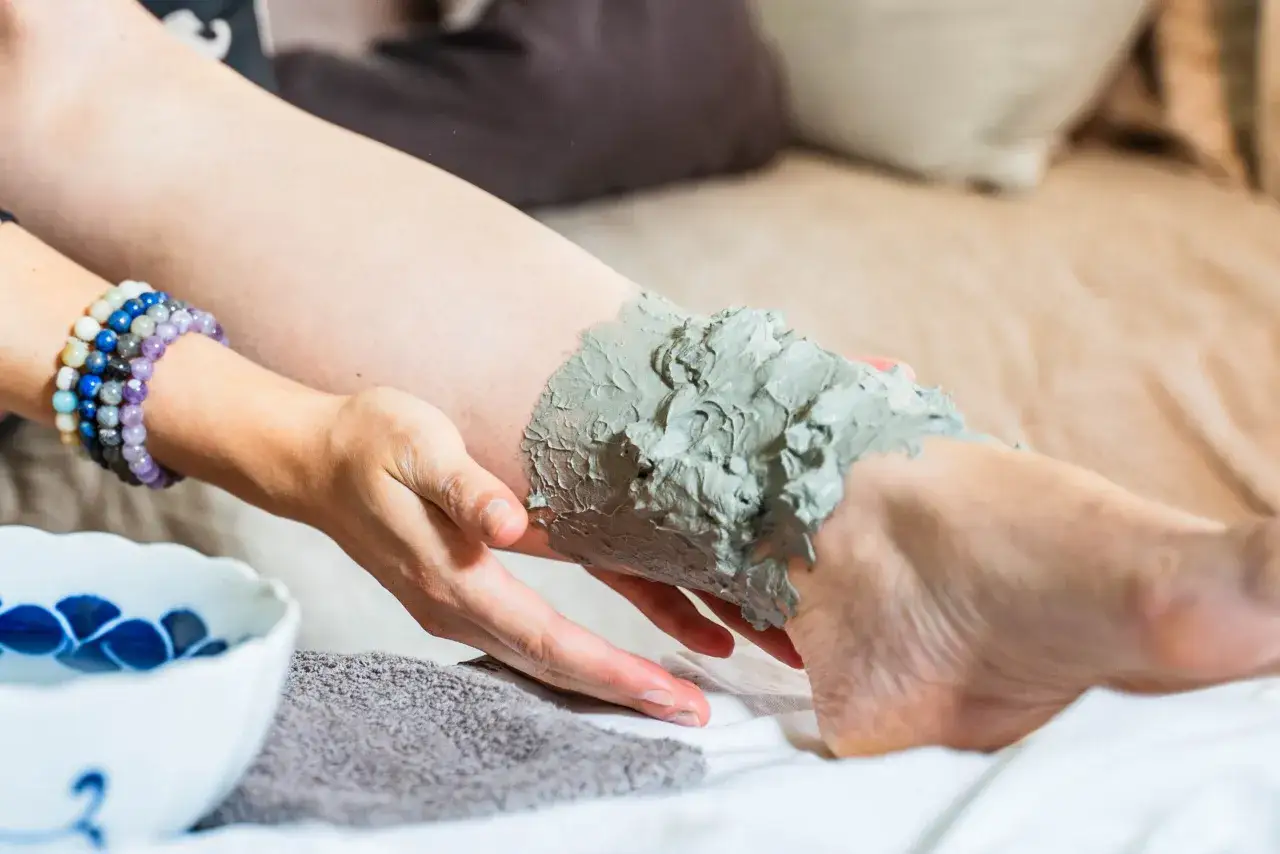 Application d'argile verte sur une jambe pour un soin détoxifiant et dégonflant. Une main applique la pâte onctueuse.