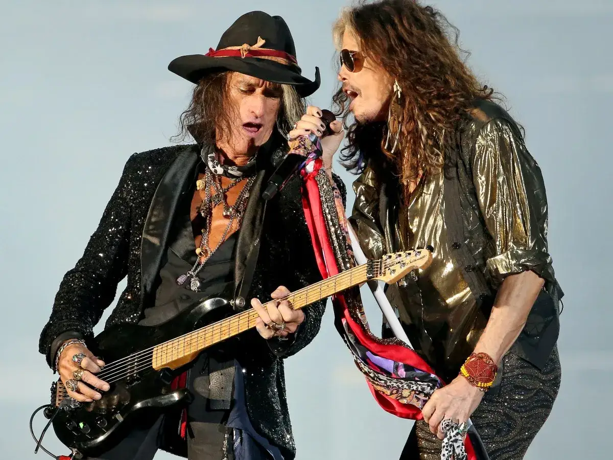 Aerosmith członkowie: Joe Perry gra na gitarze, a Steven Tyler śpiewa do mikrofonu, obaj w rockowych strojach.
