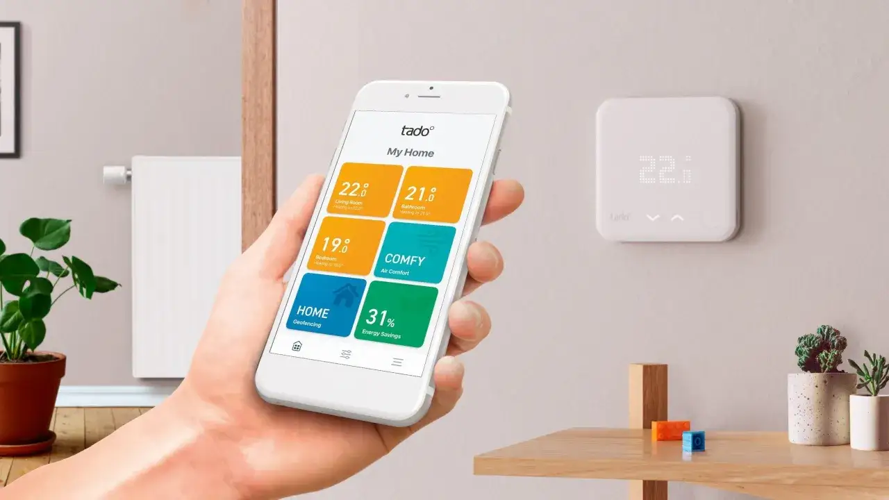 Main d'une personne tenant un smartphone affichant une application de **protocole domotique** pour contrôler le chauffage, à côté d'un thermostat mural.
