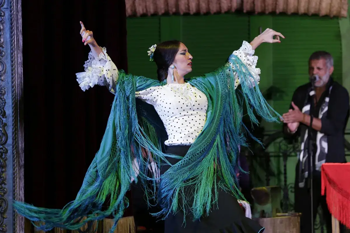 Bailarina flamenca con mantón verde, interpretando apasionadas canciones flamencas de amor, mientras un hombre aplaude al fondo.