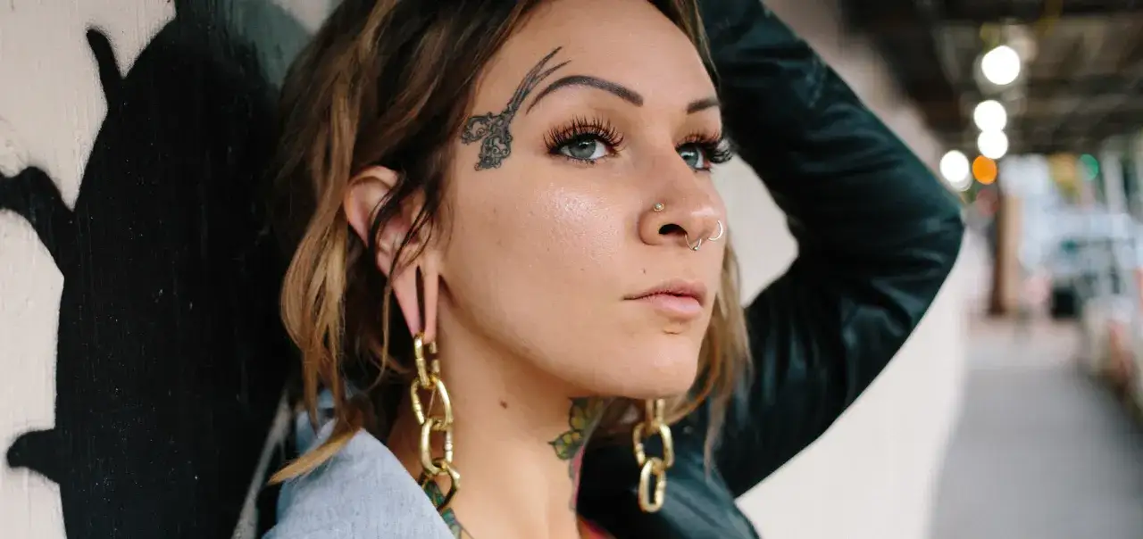 Kobieta z tatuażem nożyczek na czole i kolczykiem w nosie septum. Długie, złote kolczyki zwisają z jej uszu.