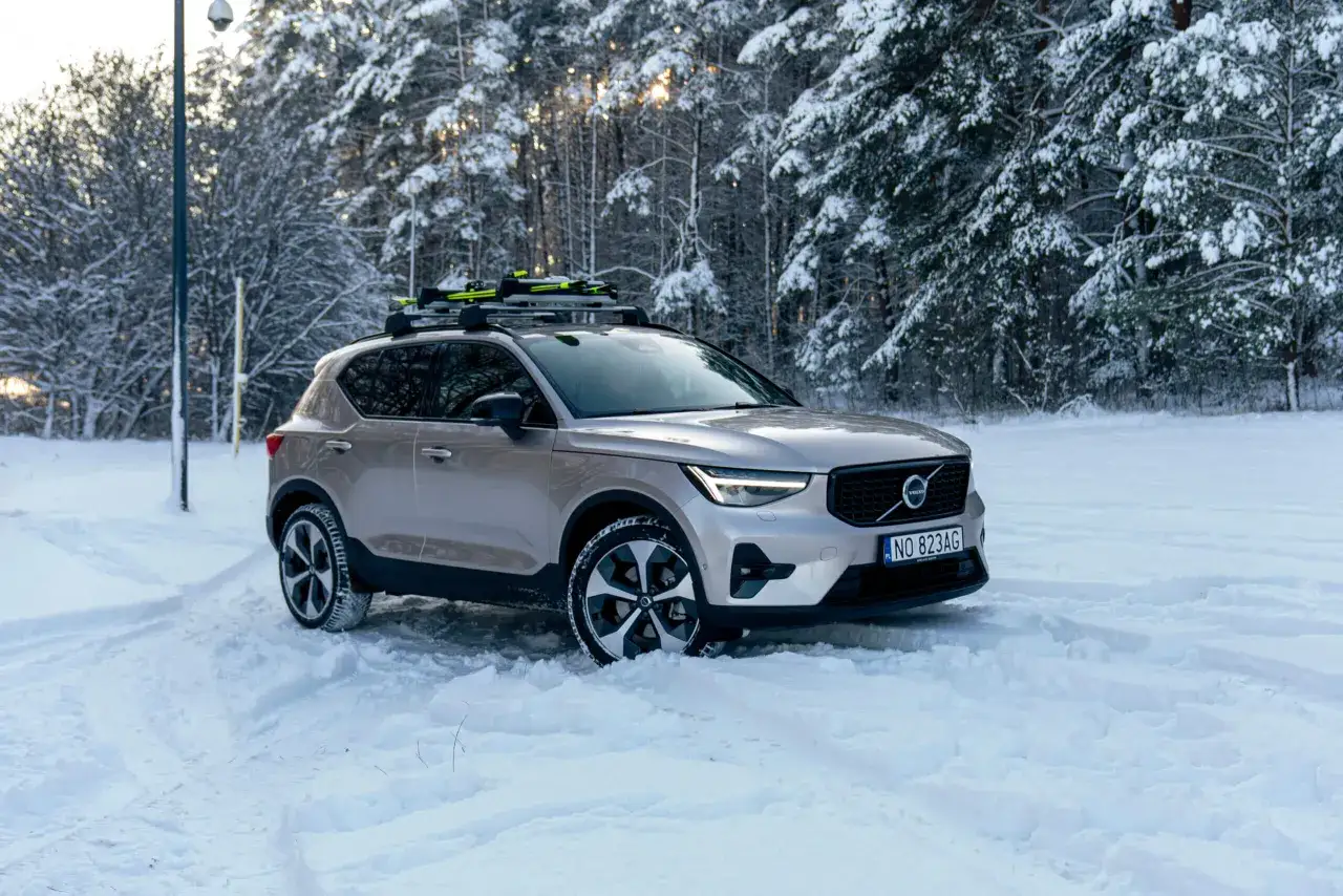 Beżowe Volvo XC40 2021 z nartami na dachu, gotowe na zimową przygodę w zaśnieżonym lesie.