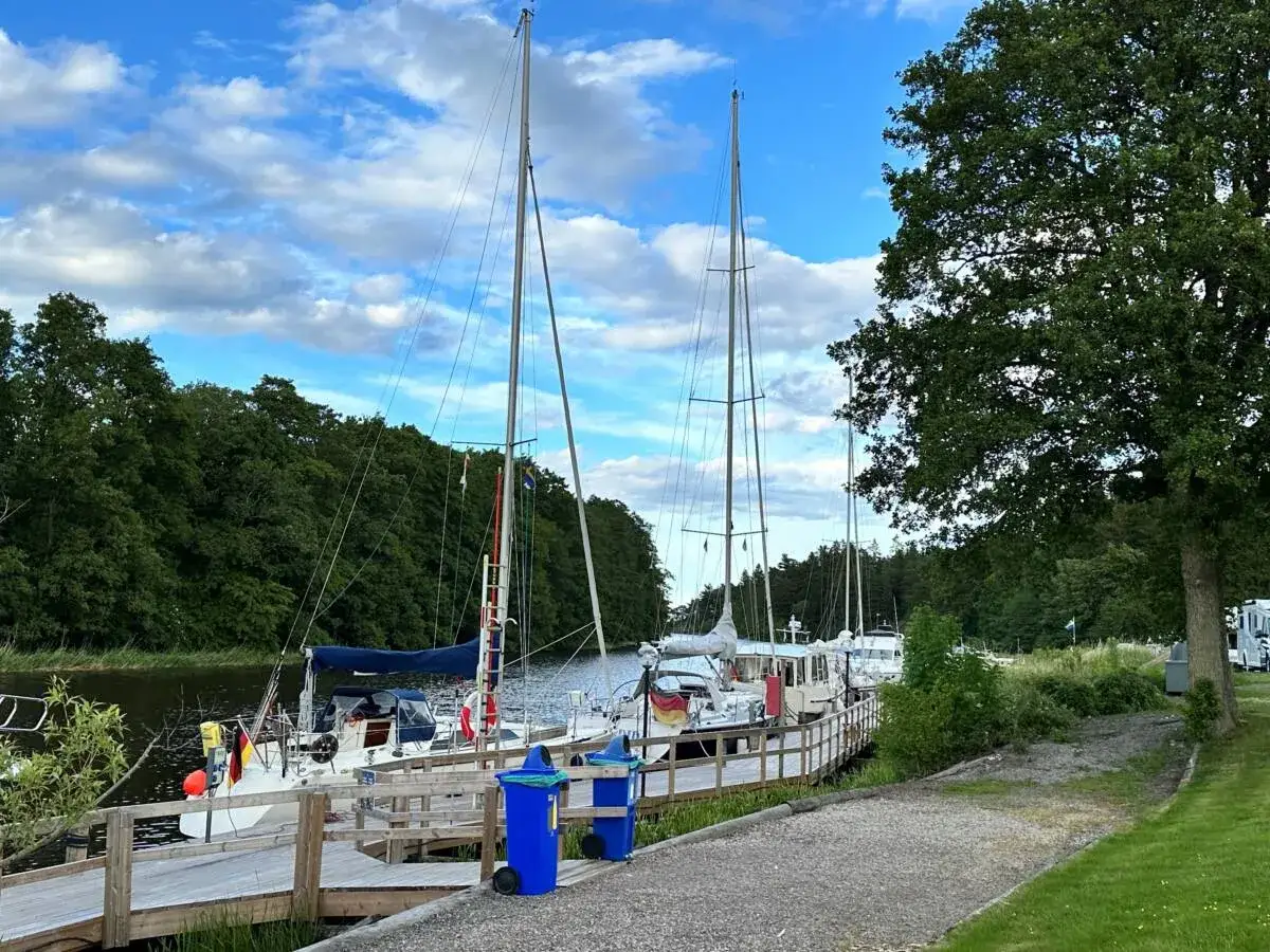Segelbåtar vid kajen på Dalbergså camping. Grönskande träd och blå himmel med moln.