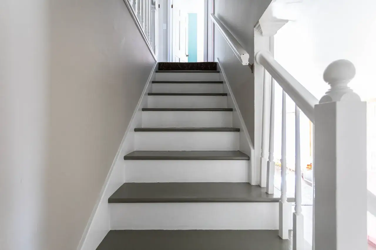 En vitmålad trappa med mörkgrå steg, redo för renovering. En ny matta på översta steget ger en känsla av pågående projekt.