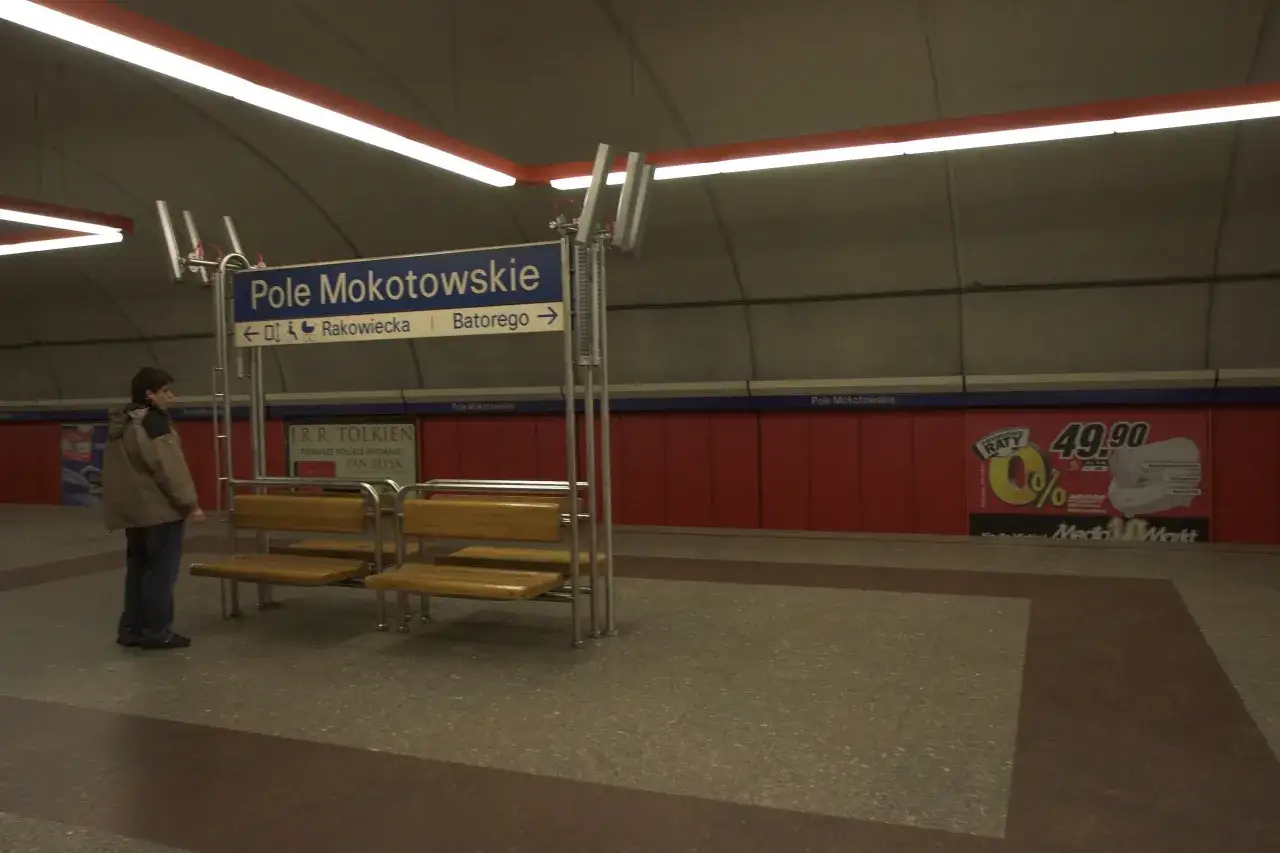 Na stacji metra Pole Mokotowskie stoi mężczyzna, obok ławki i tablicy kierunkowej.