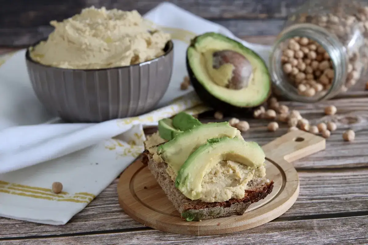 Fetta di pane integrale con hummus di avocado e fette di avocado fresco. Sullo sfondo, una ciotola di hummus, un avocado tagliato a metà e ceci in un barattolo.