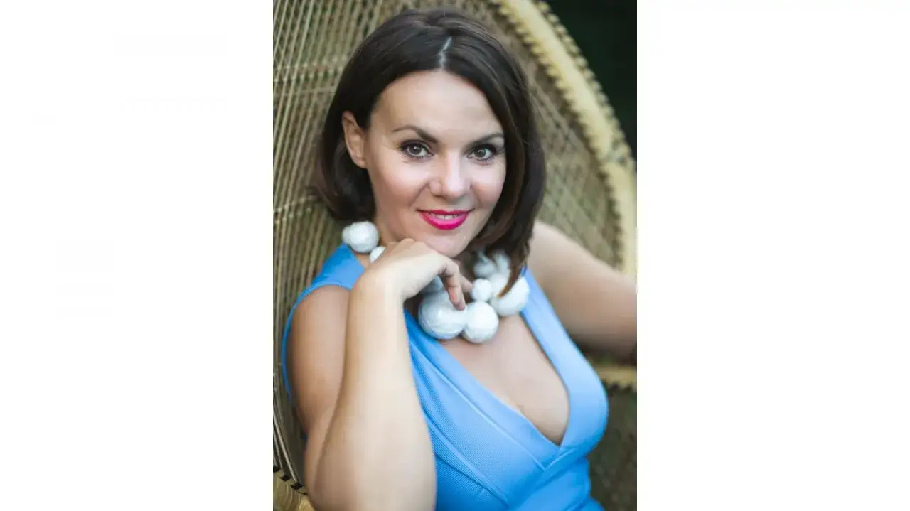 Krystyna Łuczak-Surówka, wiek nieznany, w błękitnej sukience i białym naszyjniku, siedzi w wiklinowym fotelu.