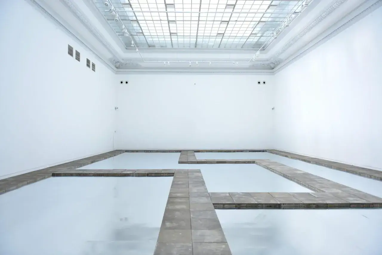 Galeria sztuki z lustrzaną podłogą i kamiennymi ścieżkami, tworzącymi labirynt. Kojący widok, jakbyś spacerował po wodzie.