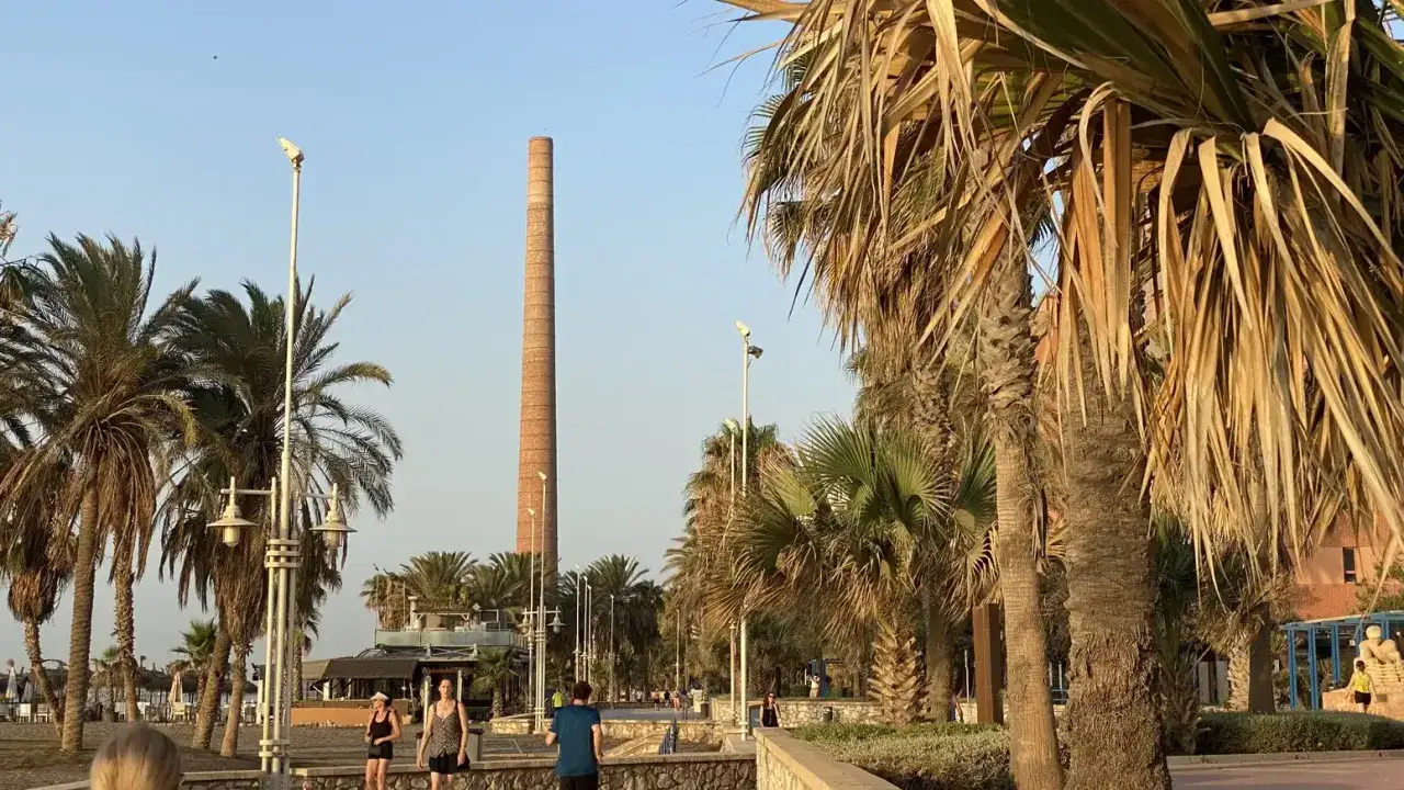 Palmen s&auml;umen den Weg am playa de la misericordia, mit einem hohen Ziegelschornstein im Hintergrund.