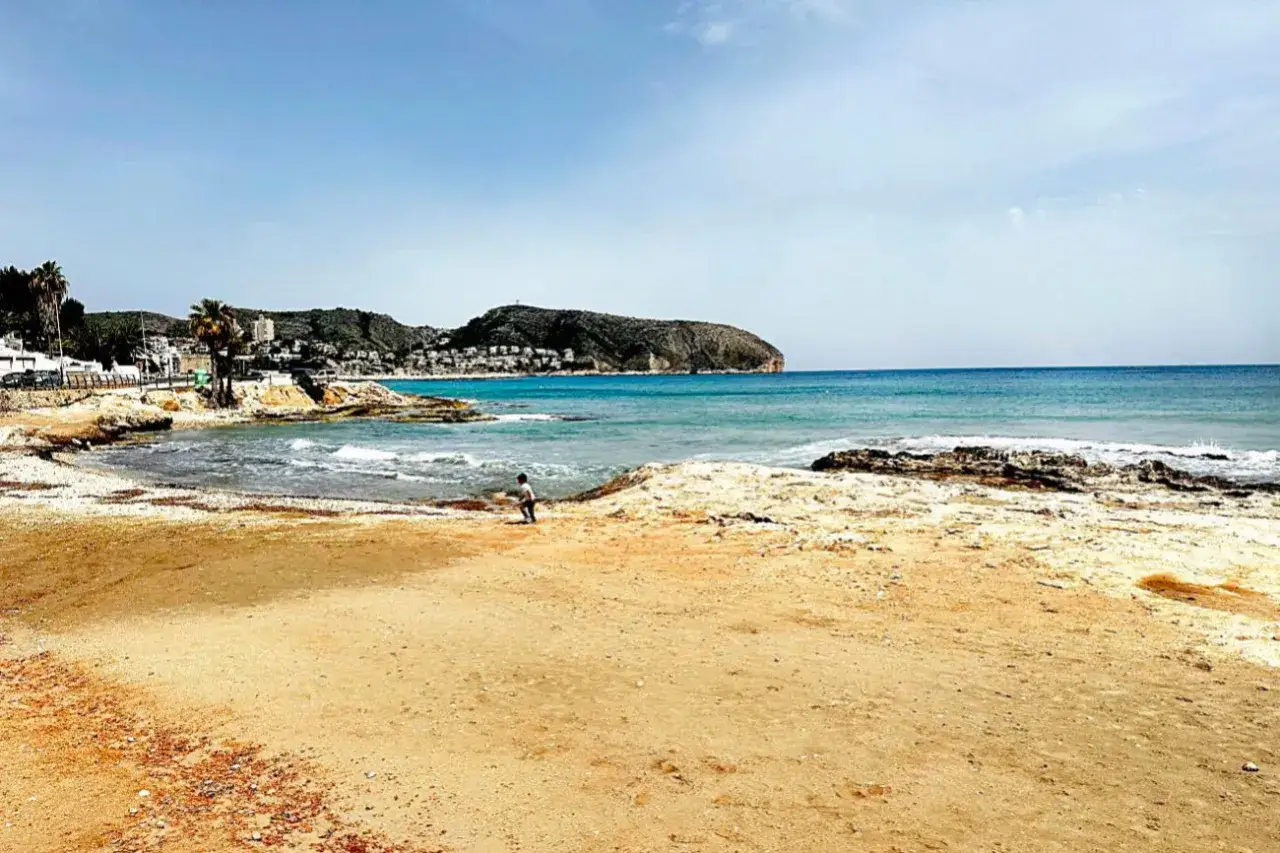 Niño juega en la arena dorada de cala l'andragó, con el mar azul y montañas al fondo.