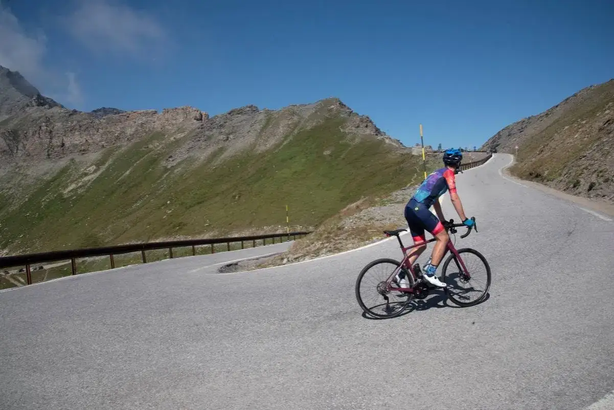 Ciclista affronta tornanti sul **colle dell'Agnello**. Montagne verdi e cielo azzurro.