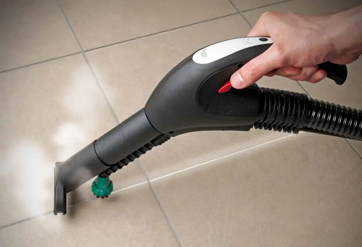 Mano che usa un pulitore a vapore per pulire le fughe delle piastrelle. Il vapore esce dall'ugello, pronto a sanificare.