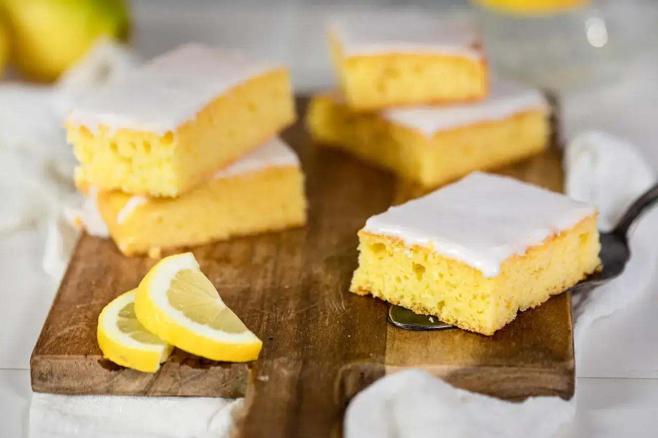 Saftiger zitronenkuchen blech, in Stücke geschnitten und mit Zuckerguss überzogen, liegt auf einem Holzbrett. Frische Zitronenscheiben garnieren die Leckerei.