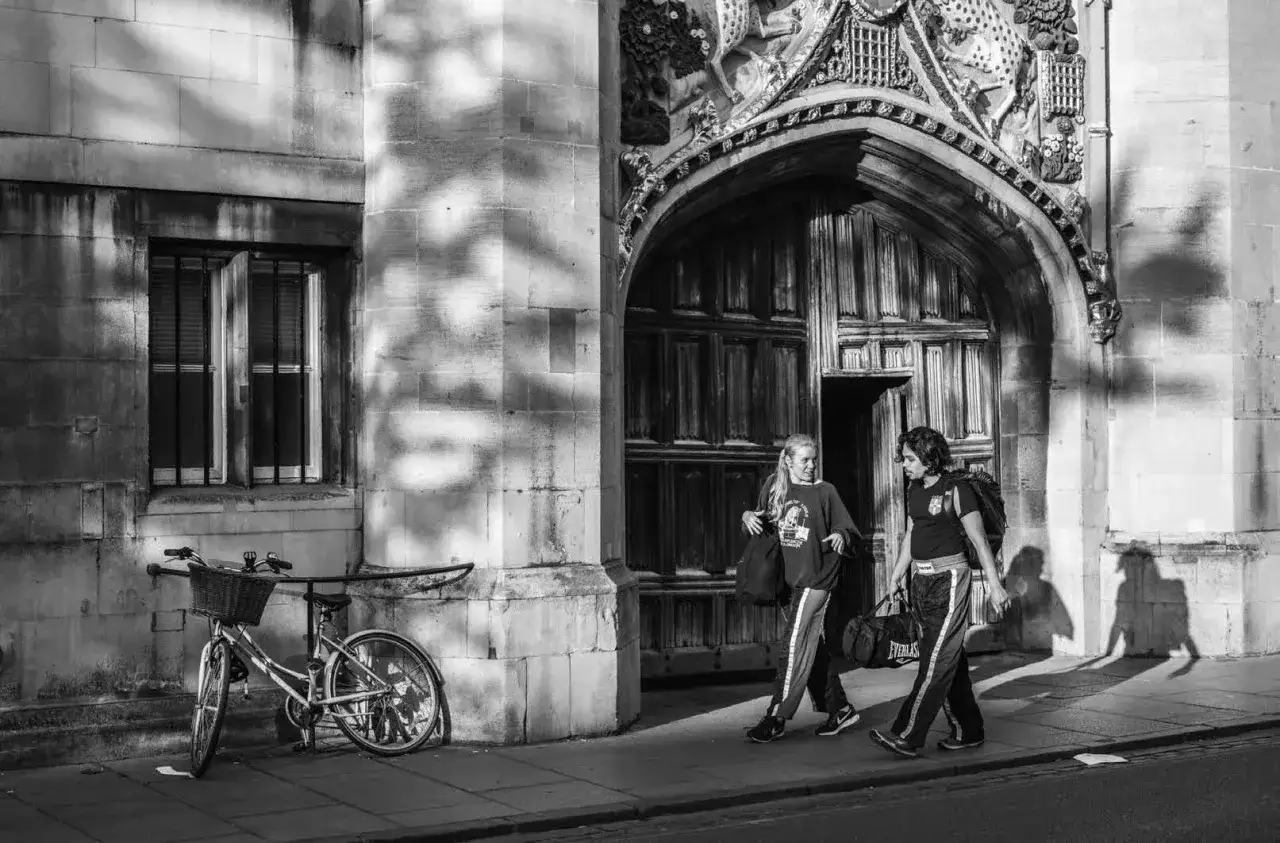 Dos j&oacute;venes caminan bajo una arcada, capturados con una Leica M11 Monochrom. Una bicicleta apoyada en la pared, sombras alargadas.
