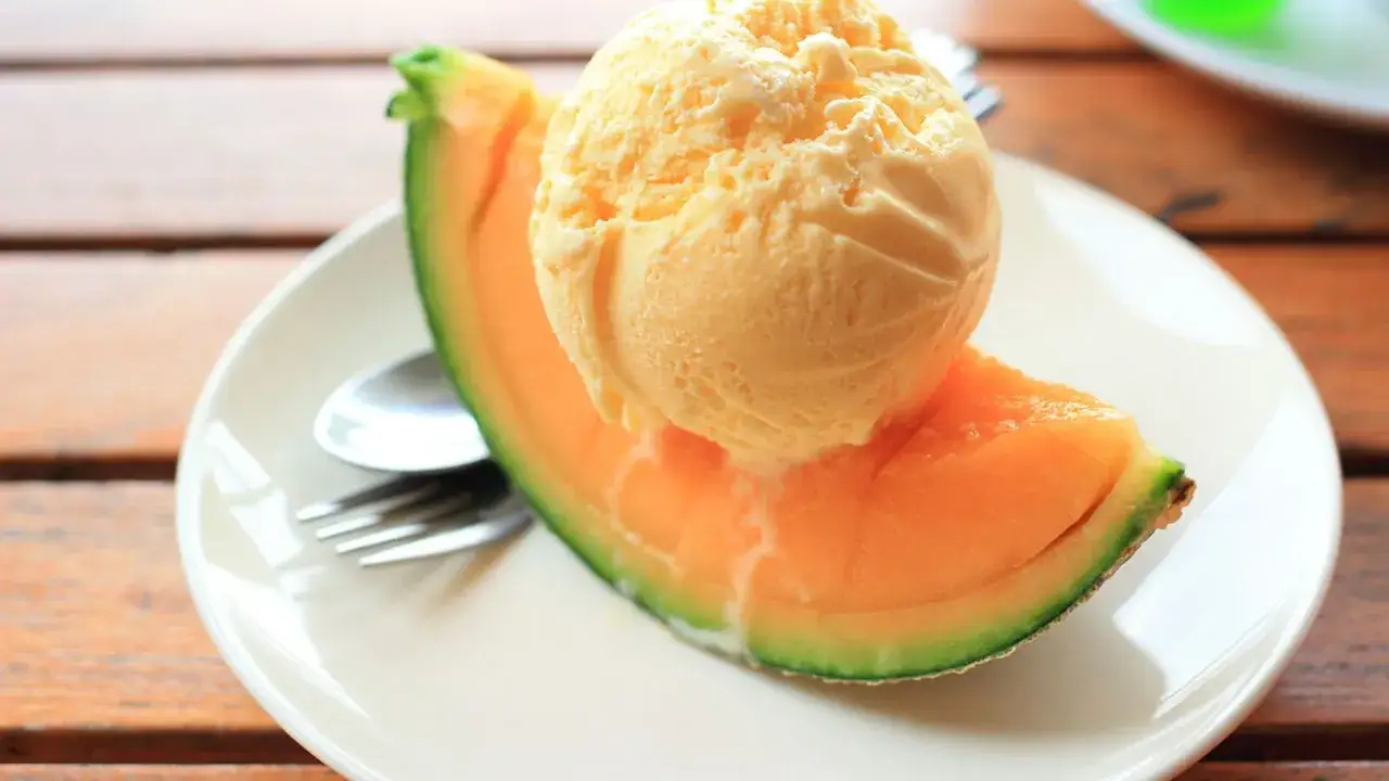 Una bola cremosa de helado de melón se derrite sobre una jugosa rebanada de melón fresco en un plato blanco.