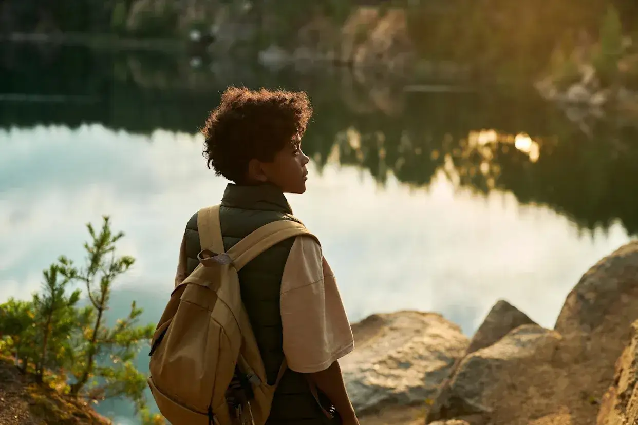 Ragazzo con zaino guarda un lago, riflettendo sul bisogno di viaggiare e sulla psicologia dell'esplorazione.