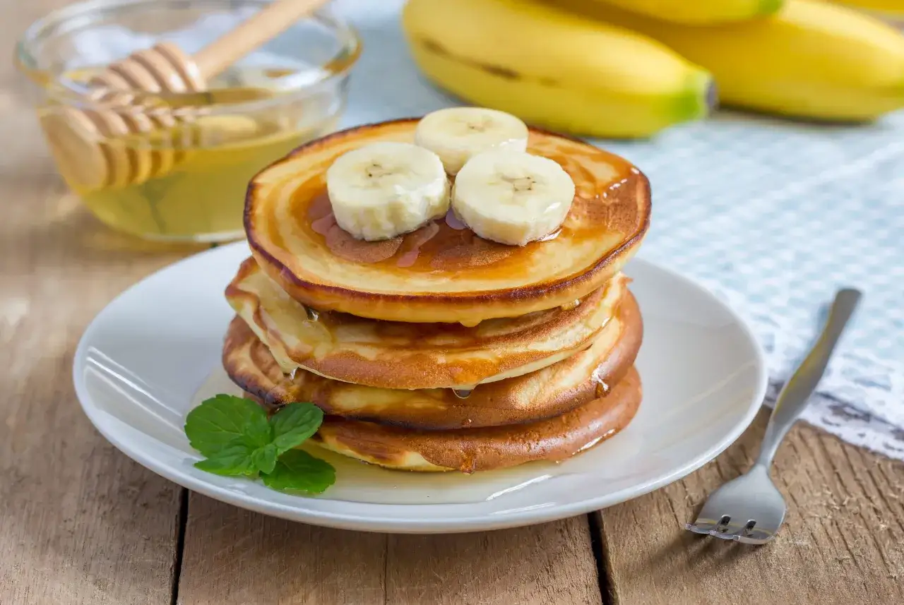 Stosik puszystych placuszk&oacute;w bananowych polanych miodem, udekorowany plasterkami banana i listkiem mięty. Obok miseczka z miodem i widelczyk.