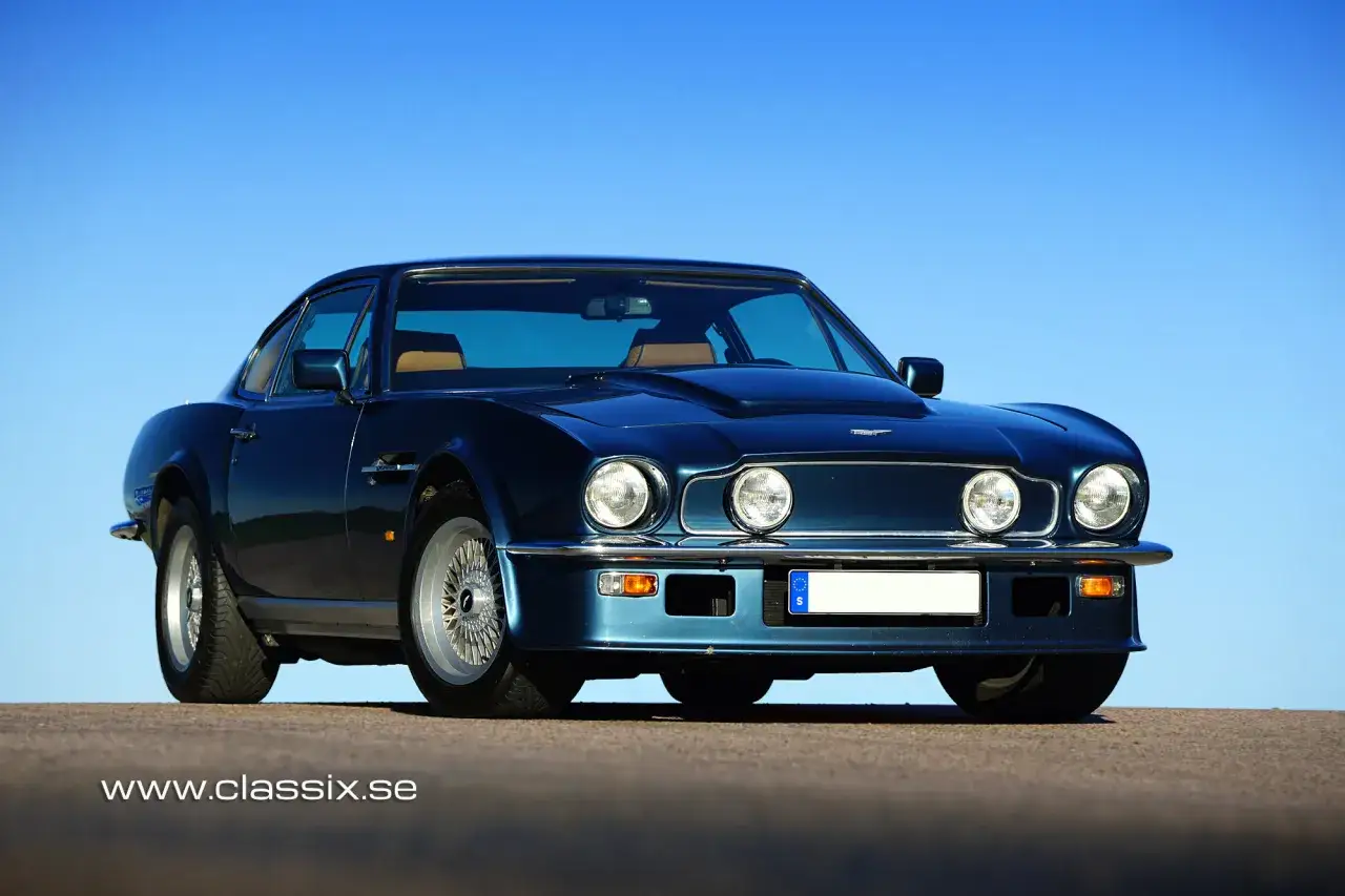En mörkblå Aston Martin V8 Vantage står parkerad mot en klarblå himmel. Denna klassiska sportbil har en tidlös design.