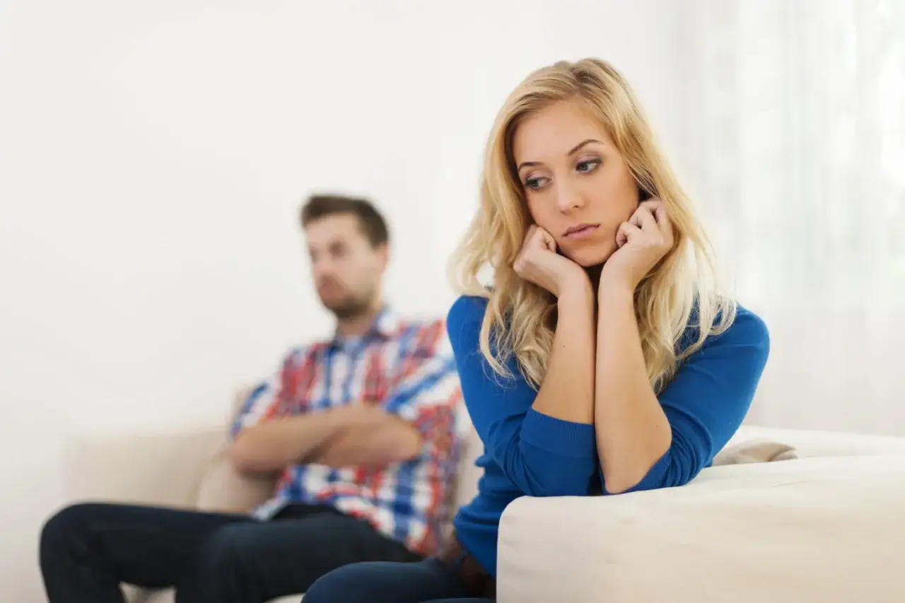 Una donna pensierosa sul divano, mentre un uomo con le braccia conserte la guarda da lontano. Un momento di tensione, quando l'amore diventa ossessione.