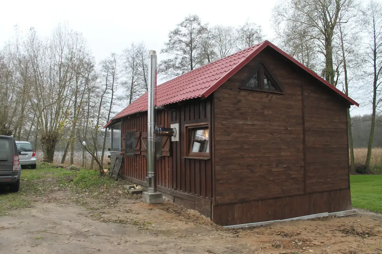 Drewniana chata z czerwonym dachem, a obok niej błyszczący, metalowy komin zewnętrzny do kozy, który zapewnia ciepło w chłodne dni.