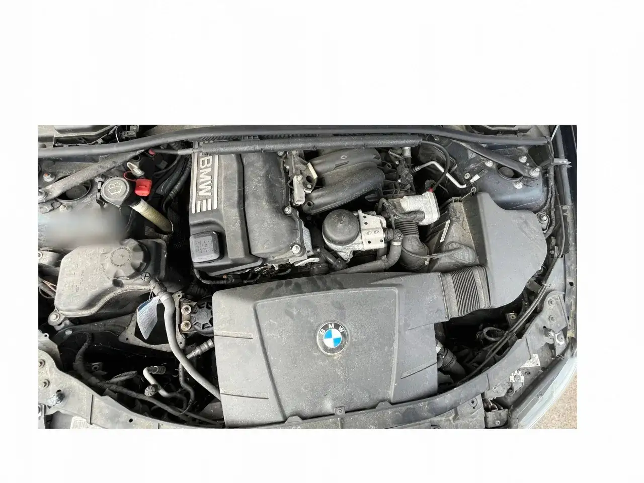 Silnik BMW E90 2.0 benzyna. Widoczny blok silnika, osłony, przewody i elementy układu dolotowego.