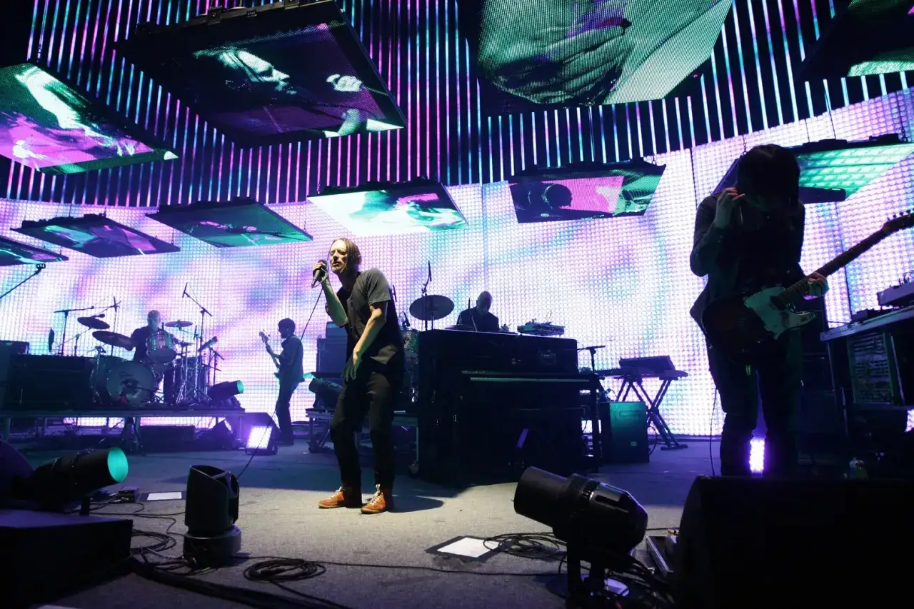 Les membres de Radiohead sur scène, éclairés par des lumières violettes et roses, avec des écrans diffusant des images.