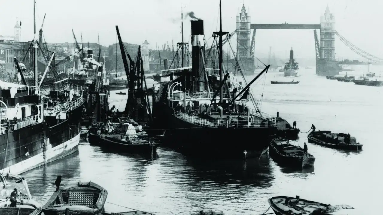 Dampfschiffe und Kräne im geschäftigen London Harbour. Im Hintergrund ragt die Tower Bridge empor.