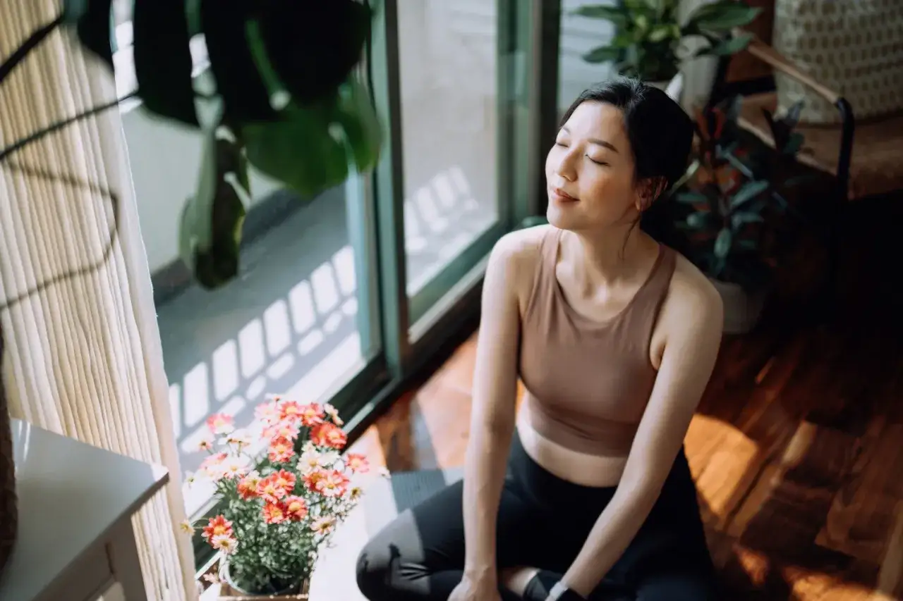 Une femme médite, profitant des bienfaits du yoga dans une pièce lumineuse. Des plantes vertes et des fleurs ajoutent à l'atmosphère paisible.