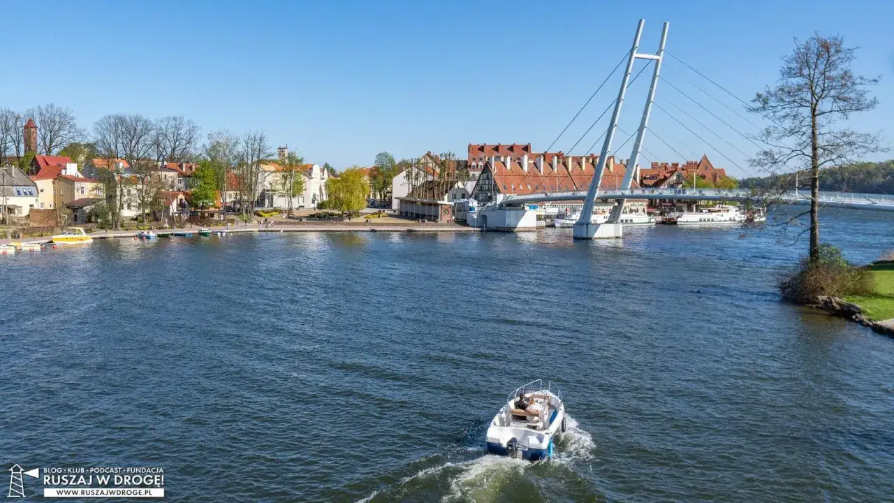 Mikołajki atrakcje: łódź motorowa płynie po jeziorze w kierunku mostu, za nim urokliwe miasteczko z czerwonymi dachami.