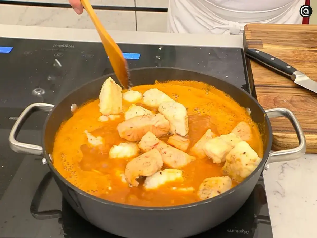Un chef remueve trozos de pescado y patatas en una cazuela con una salsa naranja, preparando una deliciosa caldereta de pescado.
