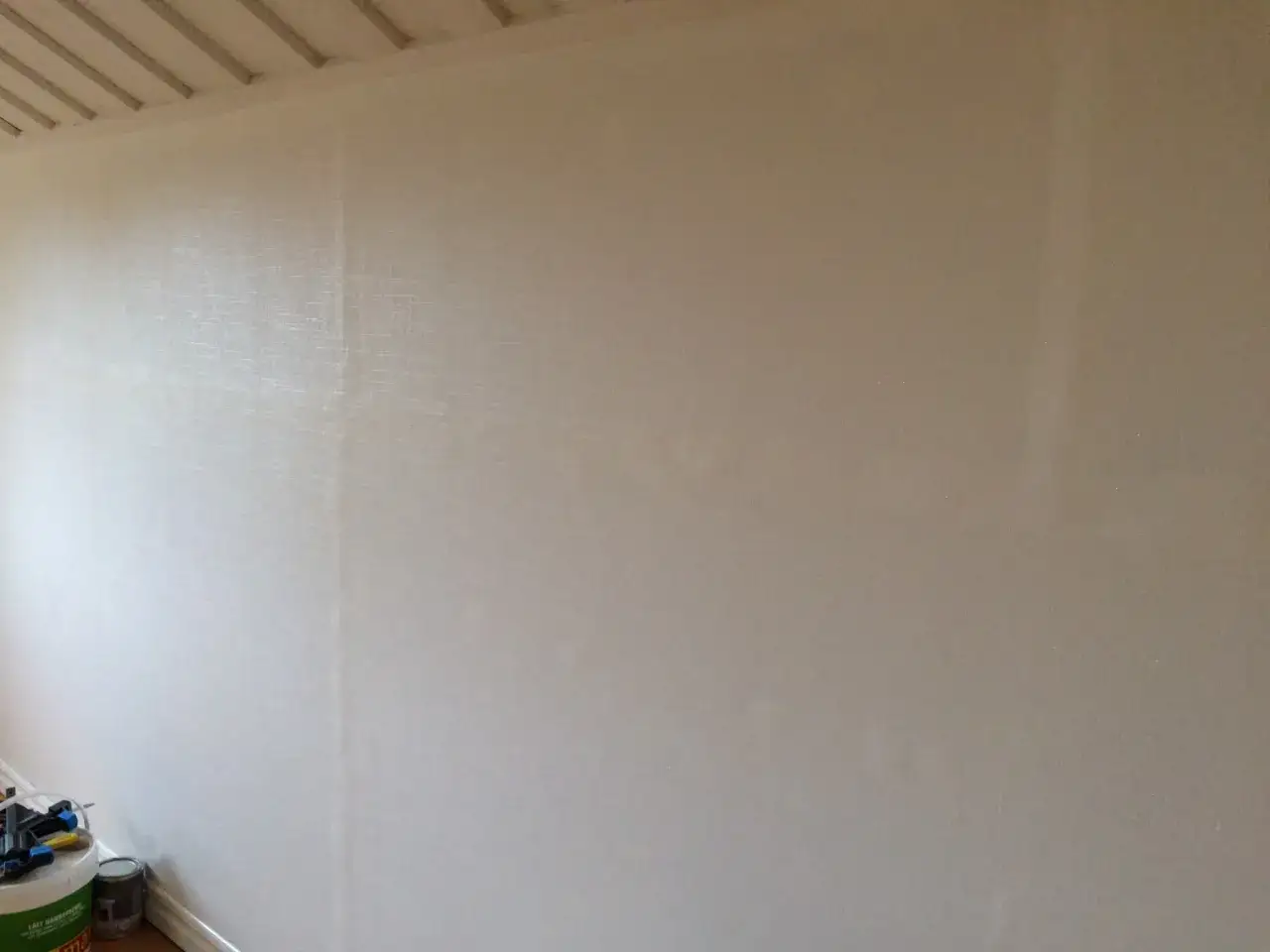 En vägg med en ljus renoveringstapet över en strukturtapet. Verktyg och färgburkar står vid golvet.