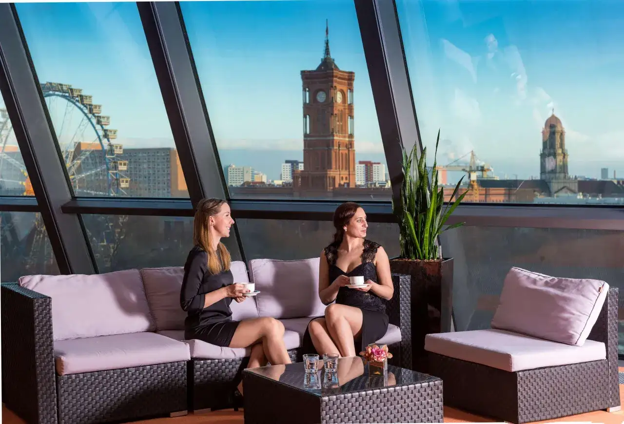 Dwie kobiety piją kawę na sofie z widokiem na Berlin. W tle widać Riesenrad i wieżę ratuszową, charakterystyczne dla **Radisson Blu Berlin**.