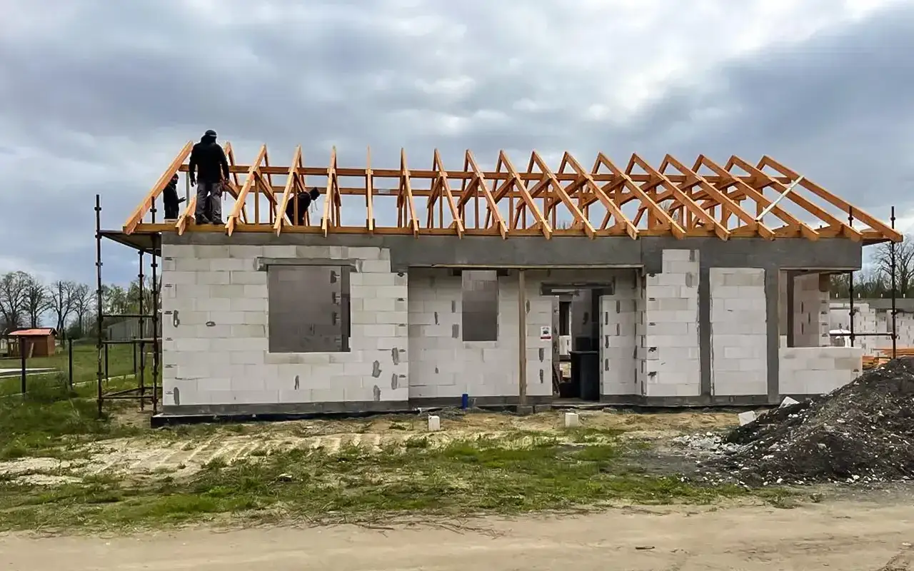 Budowa domu w toku. Pracownicy na dachu, zabezpieczeni urządzeniem samohamownym do pracy w poziomie, montują więźbę dachową.