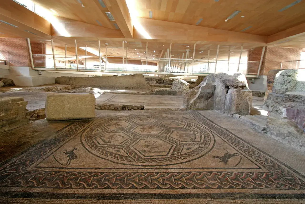 Mosaico circolare con scene di caccia e animali nella **Domus del Chirurgo**, sotto una moderna copertura in legno e vetro.