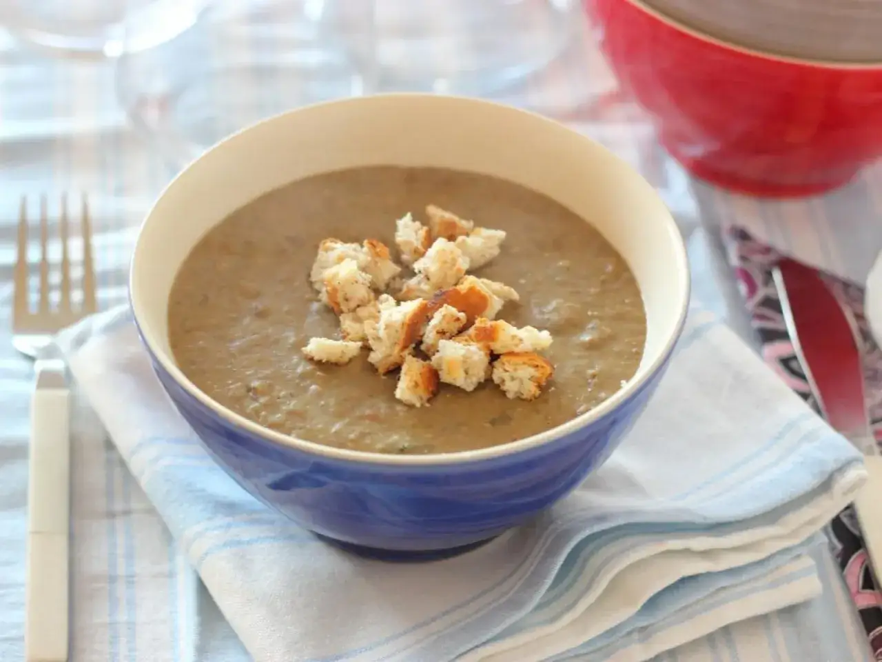 Un reconfortante plato de pur&eacute; de lentejas, coronado con crujientes picatostes, servido en un cuenco azul sobre una servilleta a rayas.