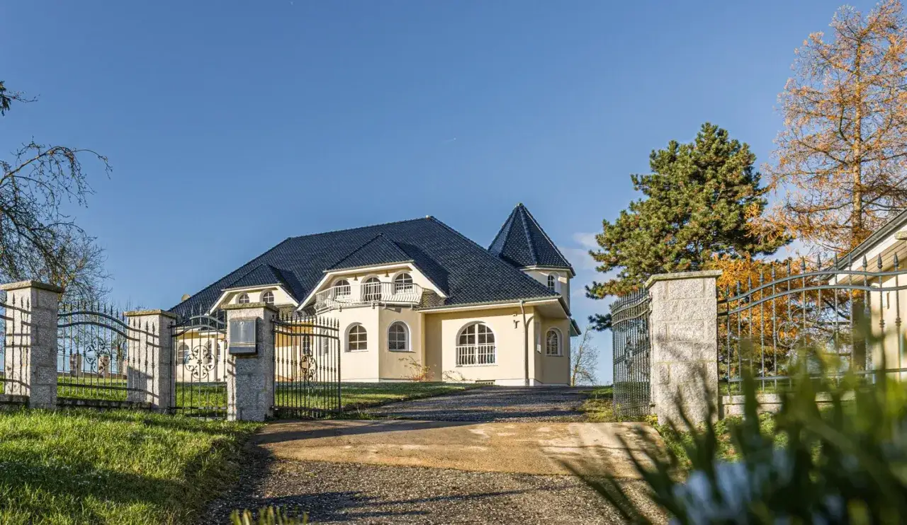 Elegantes Haus mit dunklem Dach und gelber Fassade, umgeben von einem schmiedeeisernen Tor. Ideal, um steuerfrei zu verkaufen und den Traum vom Eigenheim zu verwirklichen.