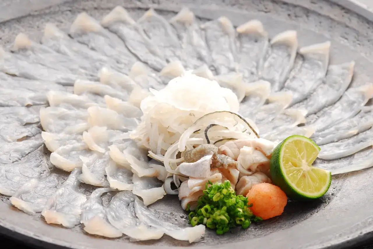 Wykwintne sashimi z ryby fugu ułożone wachlarzowo, z dodatkami: posiekanym szczypiorkiem, pomarańczową kulką i limonką.