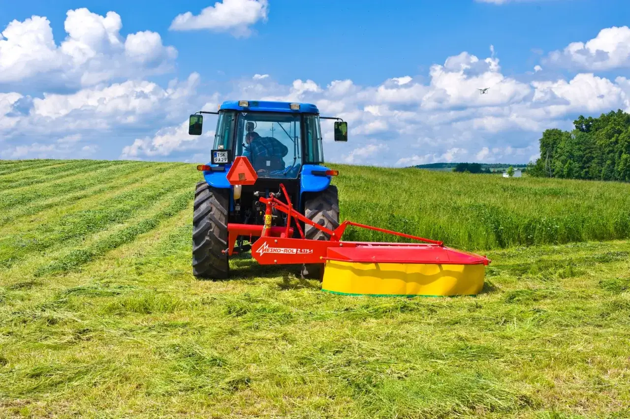 Niebieski traktor z kosiarką rotacyjną Mesko-Rol Z-175-1 pracuje na zielonym polu, tworząc r&oacute;wniutkie pasy skoszonej trawy.